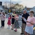 Photo seven parishoners standing outside the front of the church, facing opposite the priest and five males. Most (but not all!) of the parishoners are female, and each is holding either a candle or sheet music.