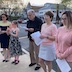 Another photo of some parishoners standing outside the front of the church, facing opposite the priest and five males. Most (but not all!) of the parishoners are female, and each is holding either a candle or sheet music.