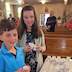 A boy and girl pose for a photo while standing in church at the front of the pews. Each is holding a cup of bread, and they're standing next to a table with blessed bread.