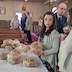 A young female is standing at a long table containing Paska breads that are wrapped in plastic, and she's holding one of the loaves as she looks towards the camera. The table has a white tablecloth and is in front of the pews on the left-hand-side of the church (as viewed from the altar). Other parishoners can be seen talking in the background. The young woman is wearing glasses.