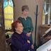 Two women in the choir loft are looking at music and singing. The woman to the left is seated, and the other woman is standing.