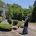 Wearing gray vestments, with a black, sleeveless top covering, Father Barnabas Fravel stands outside, at a gravesite at St. Michael's cemetery. The gravesite belongs to Fr. Dedick, a former pastor and it has a large, granite cross, surrounded by gravel, with a few shrubs, flowers, and other plants. Fr. Barnabas is holding a book and praying.