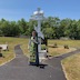 Fr. Barnabas is standing outside, in front of a large, white cross on a small pedestal, at Sts. Peter and Paul cemetery in Centralia, with his back towards the cross. He is holding a few pages of paper in his left hand. The roughly ten-foot-wide black macadam walkway on which he's standing diverges into two smaller paths that continue around either side of the the cross.