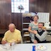 Father Barnabas is seated at a table in the church hall. A woman stands behind his right shoulder, and they both look towards the camera and smile. A gentleman in a yellow shirt and glasses is seated to Father Barnabas' right, and he is looking down as he eats.