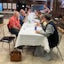 Parishioners seated at a long table that is covered with an off-white table cloth and runs from front to back in the frame. The people are eating and talking (but probably not at the same time, as that would be impolite, haha). One man, on the right-hand-side of the table is standing as he listens to a conversation.