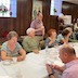 Parishioners seated at a section of a long table, which runs diagionally across the lower right corner of the frame. The people are talking and eating. A man on the far side of the table is holding a styrofoam cup while standing and listening. Another man, in the lower right corner holds a cellphone and is scrolling will talking with a woman across the table.