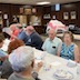 Parishioners seated at a section of a long table that runs diagionally across the lower left corner of the frame. The people are talking and eating. A man on the left side of the frame, and near side of the table, is holding a styrofoam cup while standing and listening.