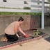 Photo two woman at our planting event. The gloved woman on the left is kneeling on black plastic in a planting bed that has brick scalloping around the edges. She's reaching underneath one of two red, trianguler pieces of fencing that will support the growing plants. The other woman is standing to her right, on the back porch. The woman on the right is holding gloves and is looking towards the camera.