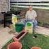 A man sitting with a bag of manure between his knees, wearing the expression that you'd expect from a man sitting with a bag of manure between his knees. Haha. Three clay pots appear in the foreground, and a green, plastic pot sits at his right side.