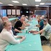 A long table, with a pastel green table cloth, runs perpendicular to the frame. Parishioners are seated at the table and conversing. A woman in a  white top is seated in the lower left corner of the frame and is looking at the camera.