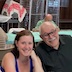 Two men and a woman smile and pose for the camera. The woman is seated between the two men who are both wearing glasses. Other tables and people appear in the background.