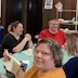 Two women seated at one of the tables look at the camera and smile. One is wearing a white top and appears in the lower left corner of the frame, while the other is wearing an orange top and appears in the lower right. Other people are visible in the background and are talking to each other.