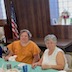 Three women are seated at the far side of a table that is covered with a pastel green table cloth. The two women on the left are looking at the camera, while the woman on the right is engaged in a conversation with someone who is off-camera.