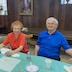 Two women and a man are seated at the far side of a table that is covered with a pastel green table cloth. The man, who appears on the right, is looking at the camera, while the two women are listening to another person who is off-camera.