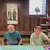 Two men are seated at the far side of a table that is covered with a pastel green table cloth. They are both looking towards the right side of the frame, listening to another person who is off-camera.