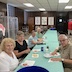 Five people are seated at a long table that runs from center frame towards the back. The table is covered with a pastel green table cloth, and the people are all looking towards the camera. People at another table are also visible in the upper right of the frame.