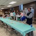 Fr. Barnabas is picking a raffle ticket from a dark, blue container. He's standing near the end of a table that runs from the lower right corner of the frame to the middle of the left side. People are seated at the far end of the table.