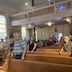 A view of the church as taken from the front of the church. Roughly twenty are standing in the pews, and a few members of the choir can be seen in the choir loft.