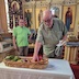 A man reaches into a wide basket of fruits and vegetables that is sitting on a table in the foreground. The table is covered by a white tablecloth, and a second man standing in the background, towards the upper left corner of the frame looks towards the first man.