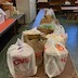 Inside what looks like a kitchen at the Mt. Carmel Food Panty, a series of folding tables run from the bottom of the frame towards the back. There are several white, plastic bags, brown, paper bags, and brown, carboard boxes on the tables, and the boxes and bags are filled with food being donated to the pantry by the church.