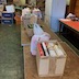Donated bags and boxes of food sit on a series of long, folding tables. The perspective of this photo is from the other end of the tables as shown in the previous photo. Empty carboard boxes are stacked against the far wall.