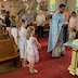 Father Aleksey, wearing a light blue vestment, is facing the right edge of the frame and reading from a paper. A row of five children of various ages are lined up to his right, with a small boy at his left. Other parishioners are also gathered around him and in the pews around him, mostly with heads bowed.