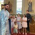 Father Aleksey, wearing a light blue vestment, is standing with his back against the left-hand frame of the photo, and he's gesturing with his right hand. A group of five children look on from his left side, which is to the right of him in the photo.