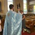 Fr. Aleksey's back fills most of the center-left frame as he blesses a woman a few feet in front of him. He is wearing a light blue vestment, and she is standing in front of a pew and wearing a gray dress with a white scarf.