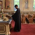 Fr. Aleksey stands in center frame, facing left and in front of a table. His head is bowed in prayer and his hands are touching the lower corners of the frame around an icon that is on the table. A plate of Prosphora bread is also on the table, to the left of the icon.