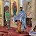Fr. Aleksey, wearing light blue vestments, stands at a podium in front of the alter, addressing the congregation. A man holding a large candle stands to his right, at the left side of the frame.