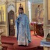 Fr. Aleksey, wearing light blue vestments, holds a cross while standing in front of the alter, addressing the congregation.