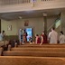 View from the front of the church, looking back as Fr. Aleksey prays while standing in the doorway between the Narthex and the Nave of the church. He has a prayer book in his hands, and parishioners in the pews are turned to face him with their heads bowed. The front door of the church is open behind him, 