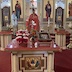 Fr. Aleksey, wearing red vestments, stands at a lecturn in front of the altar. A man, who is wearing a light blue shirt and holding a large candle, stands to Fr. Aleksey's right, towards the left-hand side of the frame. There's a table in front of Fr. Aleksey that has a red tablecloth under glass. On top of the table, from left to right, is a gold cross laying in a bed of red and white flowers, a silver cross standing vertical, and an icon on a pedestal.