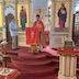 A closer-in view of Fr. Aleksey, wearing red vestments and standing at a lecturn in front of the altar. A man, who is wearing a light blue shirt and holding a large candle, stands to Fr. Aleksey's right, towards the left-hand side of the frame.