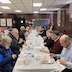 Photo from the luncheon. A long table, covered in a white tablecloth runs perpendicular from the center of the photo to the back wall. Attendees are seated along both sides of the table and are eating. A woman on the right, a third of the way down the table, is looking at the camera, but everyone else is eating or engaged in converstation. Part a second table is visible in the middle-left part of the frame, with a few people seen there.