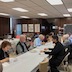 Photo from the luncheon.  View of roughly half of a long table running from the lower left corner and extending to the right-hand third of the frame. The table is covered by a white tablecloth, and people are seated on both sides of the table while eating and/or talking.