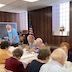 Photo from the luncheon. View of the far end of the table, which extends from the lower right corner of the frame to roughly a third of the way up the left side. The table is covered with a white tablecloth, and people are seated on either side while eating and talking. The four people visible on the left (or near) side have their backs facing the camera. Five people are shown on the far side, with a man at the far end (on the left) standing.