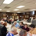 Photo from the luncheon. Somewhat wide view of the church hall showing two tables of parishioners sitting at the tables while talking and eating. A few people are standing, and a woman at the left edge of the frame appears to be looking at something on the wall that is out of frame.