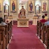 Father Timothy, wearing bronze-colored vestments, is speaking to the congregation and has his hands raised upwards. The view is from the back of the church, looking up the center aisle, and pews are visible on both sides of the frame.