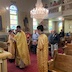 Father Aleksey stands before an icon table, holding a book and reading prayers. There is an altar boy on either side of him, and his back is facing the parishoners, many of which are visible in the background.