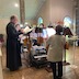 A view from the other side of the choir loft. The entire choir, consisting of five women and three men, are seen either standing or sitting in front of music stands as they sing.