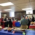 A view of the church hall where clergy and parishoners are standing, each holding a sheet of paper, and praying before the meal. There's food sitting on two tables that are perpendicular to one another. One table runs horizontally across the bottom of the frame and has a blue tablecloth, and the other runs across the right edge of the frame and has a red tablecloth.