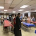 A wide view of the hall, showing three tables running from the front of the frame towards the back. The two outter tables have red tablecloths, and the center tablecloth is blue. Parishoners are standing on either side along the length of the tables.