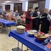 Another wide view of the hall, showing tables running from the front of the frame towards the back. The tables have red and blue tablecloths, altering by row, and parishoners stand on either side along the length of the tables. This photo combines parts of the previous two photos, as the room is a bit large to fit everything into one image.