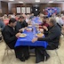 A view of the center table, which is covered by a blue tablecloth. There are clergy seated at the foreground end of the table and parishoners at the back half. They are all eating and/or talking.