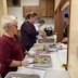 A view from the kitchen, where three woman are serving food through two open windows that open out to the main hall. There are aluminum pans of food on the counter between the women and the windows, and parts of two people are visible through the window.