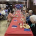 A view of the right-most table, which is covered by a red tablecloth. Parishoners are seated on both sides of the table, and they are all eating and/or talking.