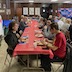 A view of the left-most table, which is covered by a red tablecloth. Parishoners are seated on both sides of the table, and they are all eating and/or talking.