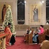 In the body of the church, Fr. Aleksey addresses several children, telling them the story of St. Nicholas. The view is from the left row of pews, with the children seated in the right-hand pews, facing towards Father Aleksey, who is seated on the left side of the frame. There's a decorated Christmas tree to his left, and in the photo, the tree appears behind him. The children are seated in the first two pews, and there's an adult male seated behind them, in the fourth pew.