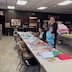 Photo of Matushka Natallia standing on the far side of a long table in the church hall. A young woman is seated at the table, to Matushka's right, which Matushka has an embracing arm around a young girl who standing to her left. The brown folding table is covered in a long sheet of white paper, with art supplies and finished Christmas cards spread across the table.